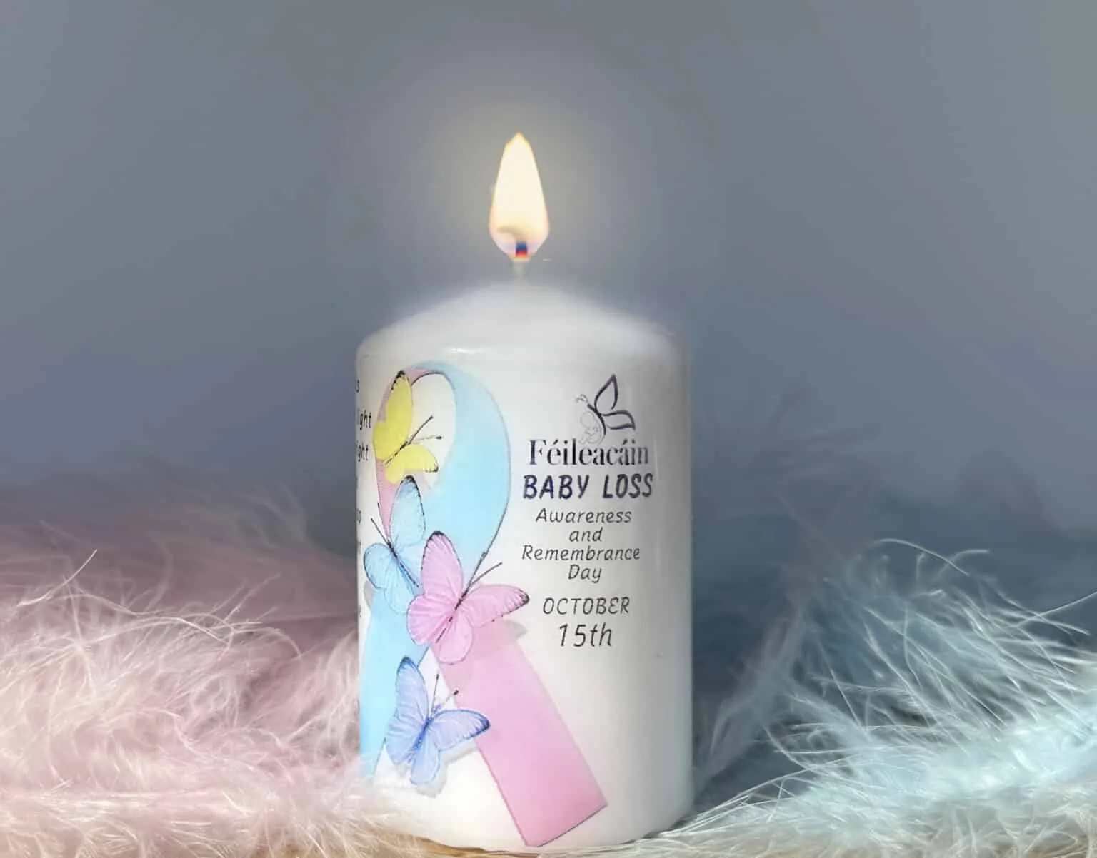 A lit white Féileacáin remembrance candle decorated with a pink and blue ribbon and butterflies, reading Baby Loss Awareness and Remembrance Day, October 15th, resting on soft pink and blue feathers