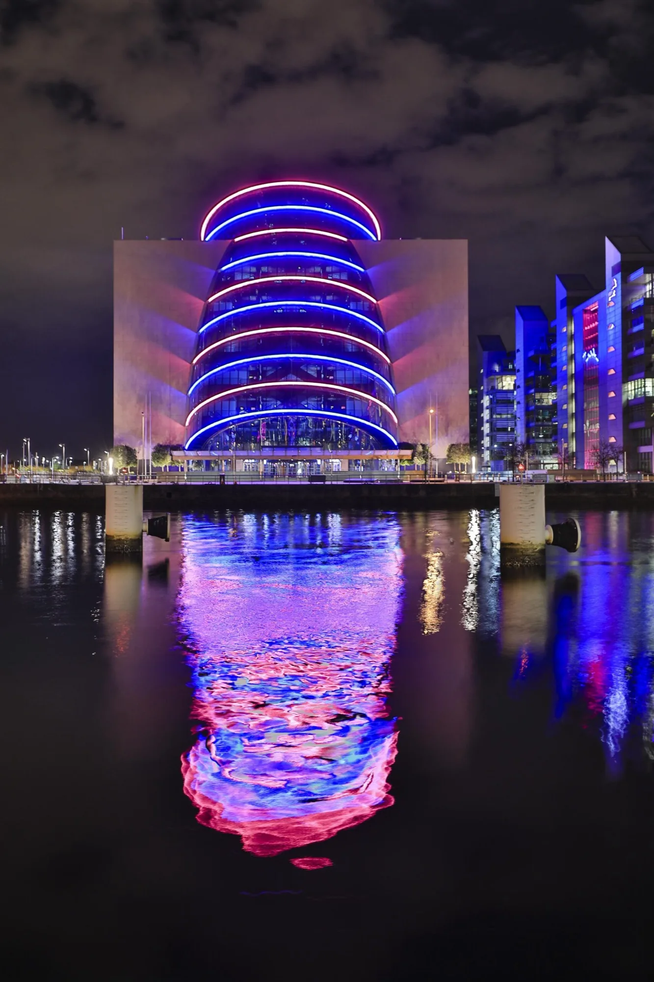 The Convention Centre Dublin at night, ringed with blue and pink light, reflected in the river below