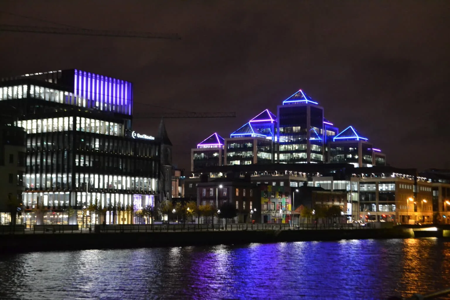 Office buildings along the river in Dublin at night, their rooftops outlined in blue and purple light